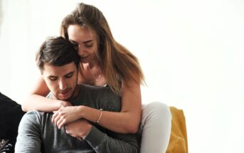 A woman with long hair affectionately hugs a man from behind as they sit together. Both are dressed in casual clothing, and the background is minimalist, focusing on their tender interaction. The mood is calm and intimate.