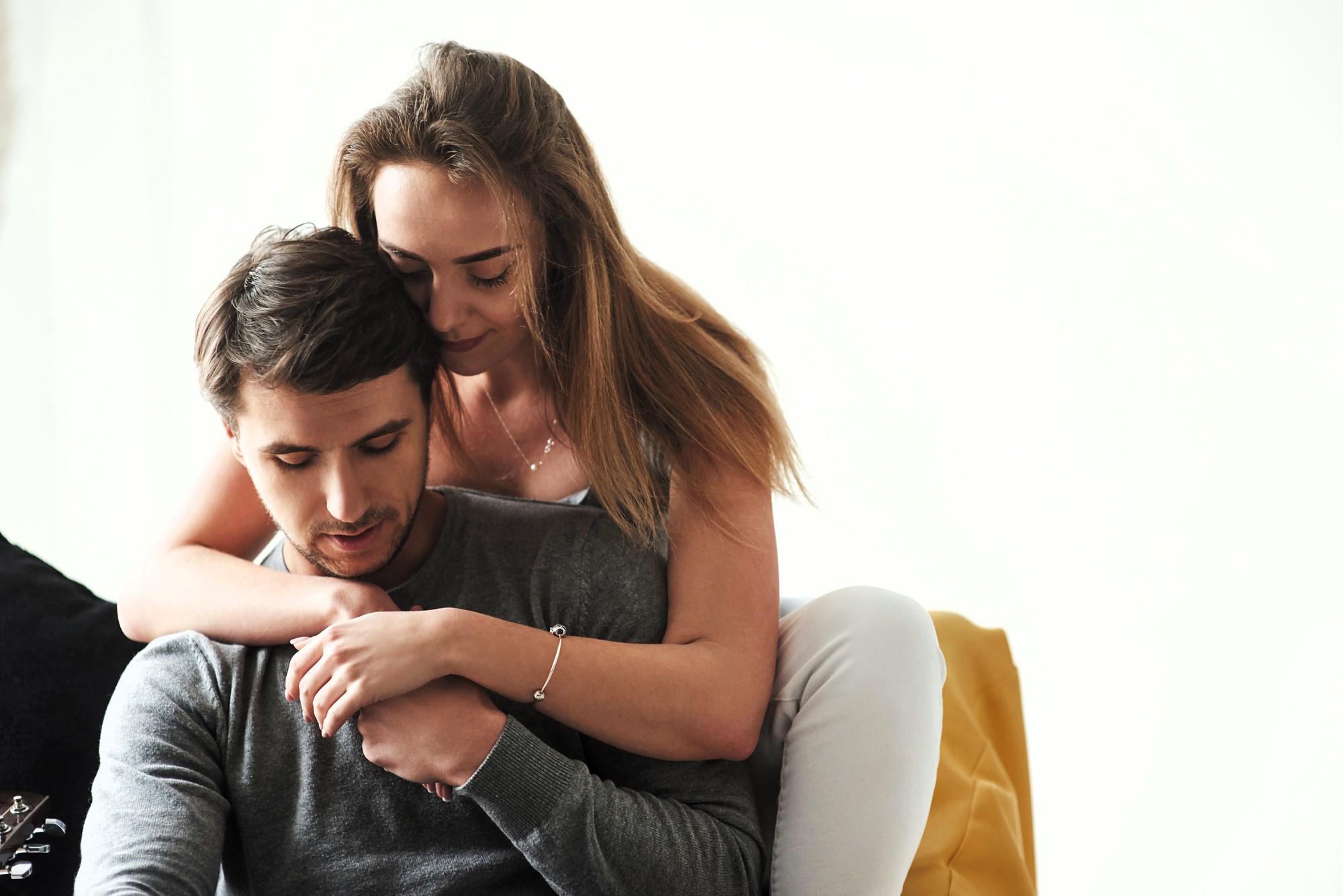 A woman with long hair affectionately hugs a man from behind as they sit together. Both are dressed in casual clothing, and the background is minimalist, focusing on their tender interaction. The mood is calm and intimate.