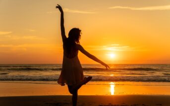 Silhouette of a person gracefully dancing on a beach at sunset, with one arm extended upward and the other at the side. The ocean waves gently lap the shore, and the sky is a gradient of orange and yellow hues.