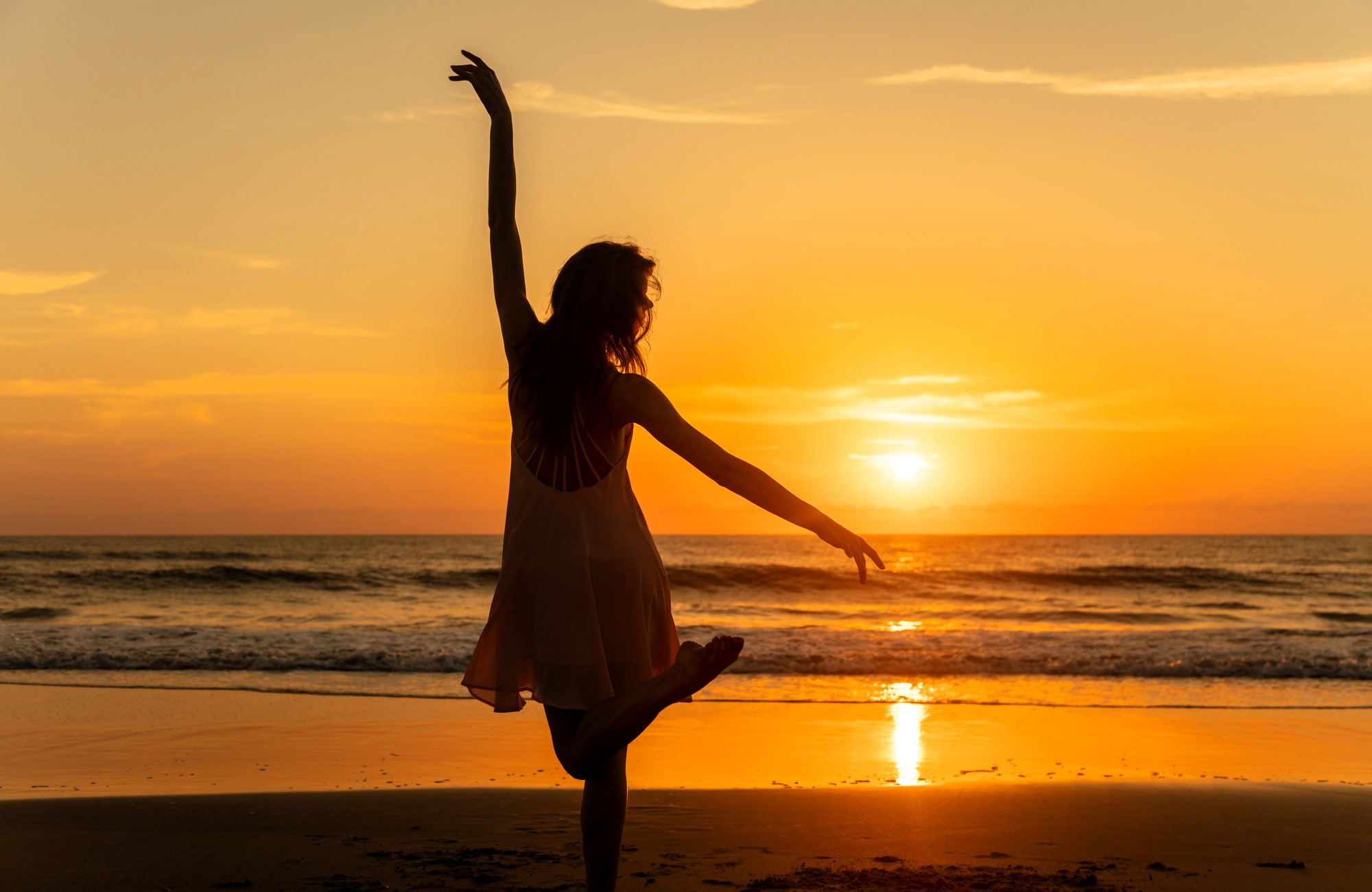 Silhouette of a person gracefully dancing on a beach at sunset, with one arm extended upward and the other at the side. The ocean waves gently lap the shore, and the sky is a gradient of orange and yellow hues.