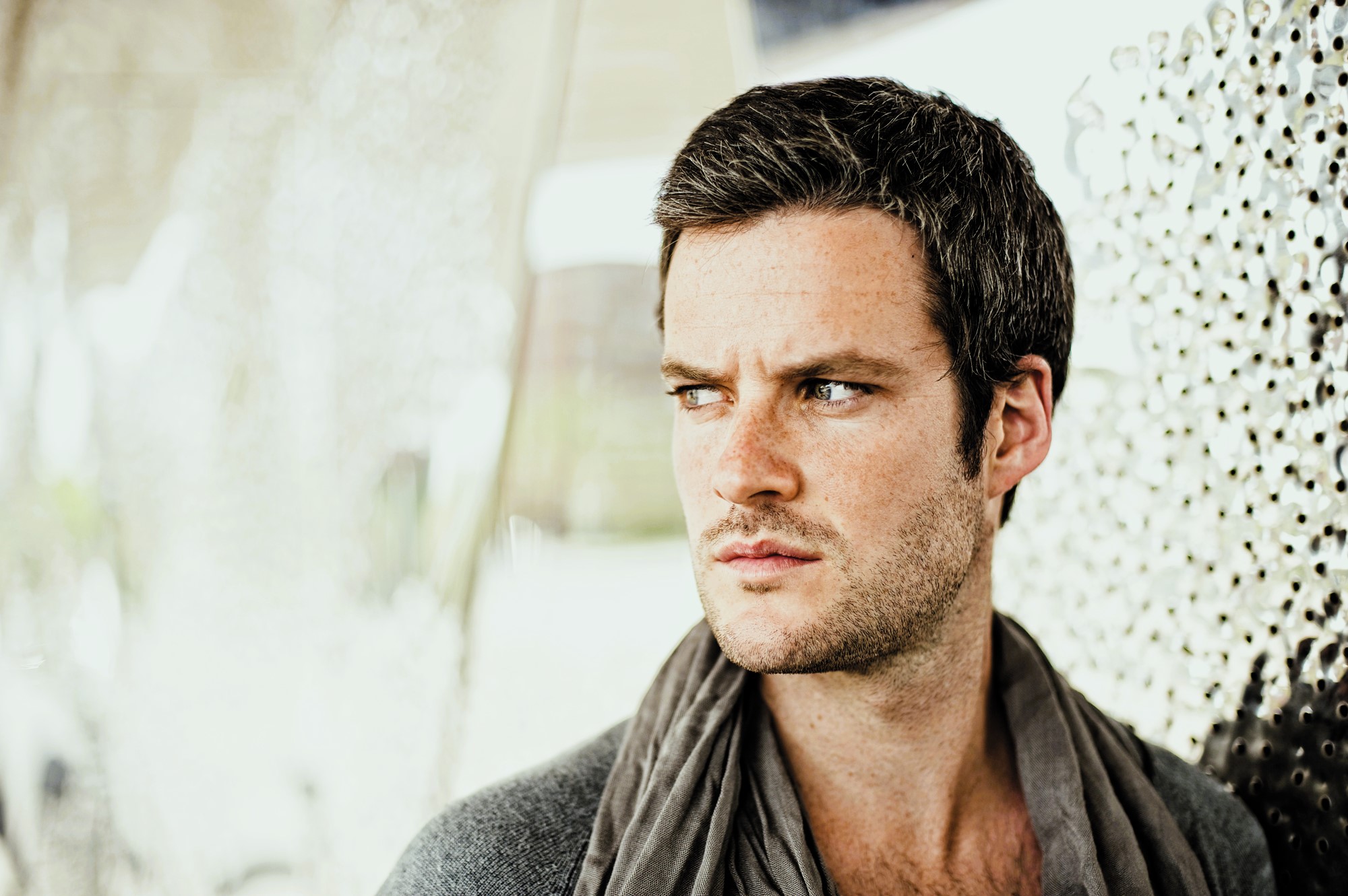 A man with short dark hair and a beard looks contemplatively to the side. He is wearing a scarf and a sweater. The background is softly blurred with abstract textures.