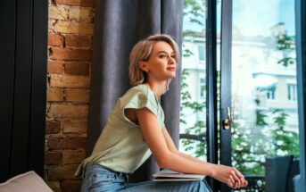 A woman with short, blonde hair sits by a window, looking outside. She is wearing a light green top and jeans, holding a closed book on her lap. The room has a brick wall and curtain, with a view of trees and buildings outside.