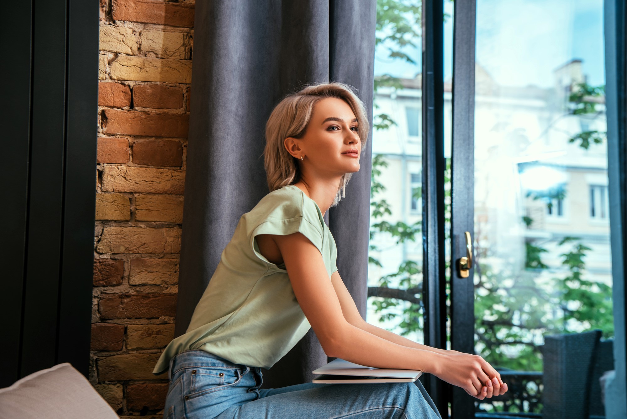 A woman with short, blonde hair sits by a window, looking outside. She is wearing a light green top and jeans, holding a closed book on her lap. The room has a brick wall and curtain, with a view of trees and buildings outside.