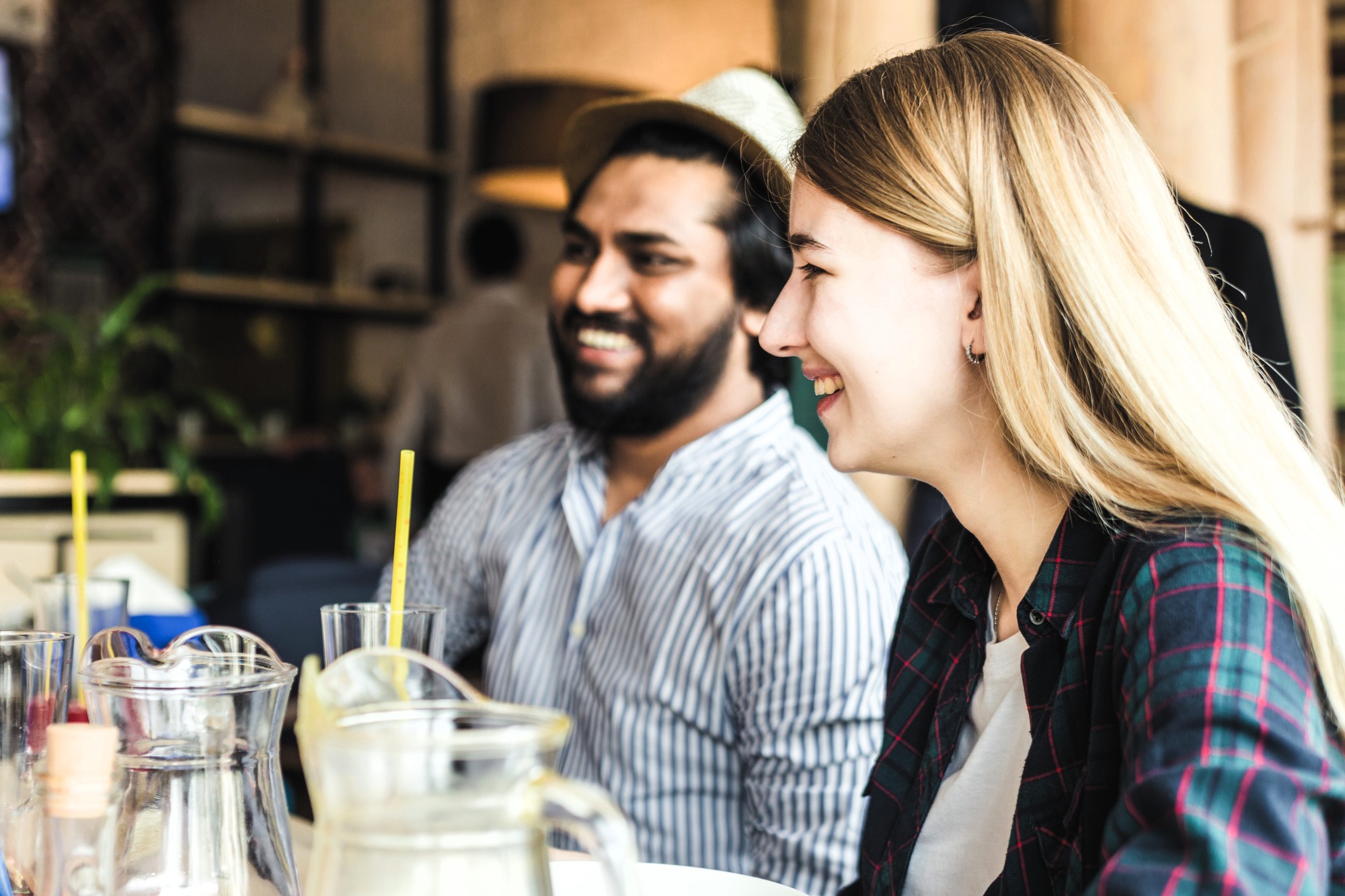 Two people sit at a table, smiling and enjoying their conversation. The person on the left wears a striped shirt and hat, while the person on the right has long blonde hair. Glasses and a pitcher are visible on the table.