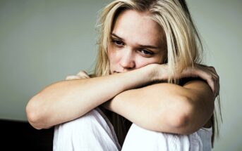 A woman with long blonde hair sits with her arms resting on her knees, looking contemplatively into the distance. She appears to be in a relaxed indoor setting, wearing a white outfit. The background is blurred.