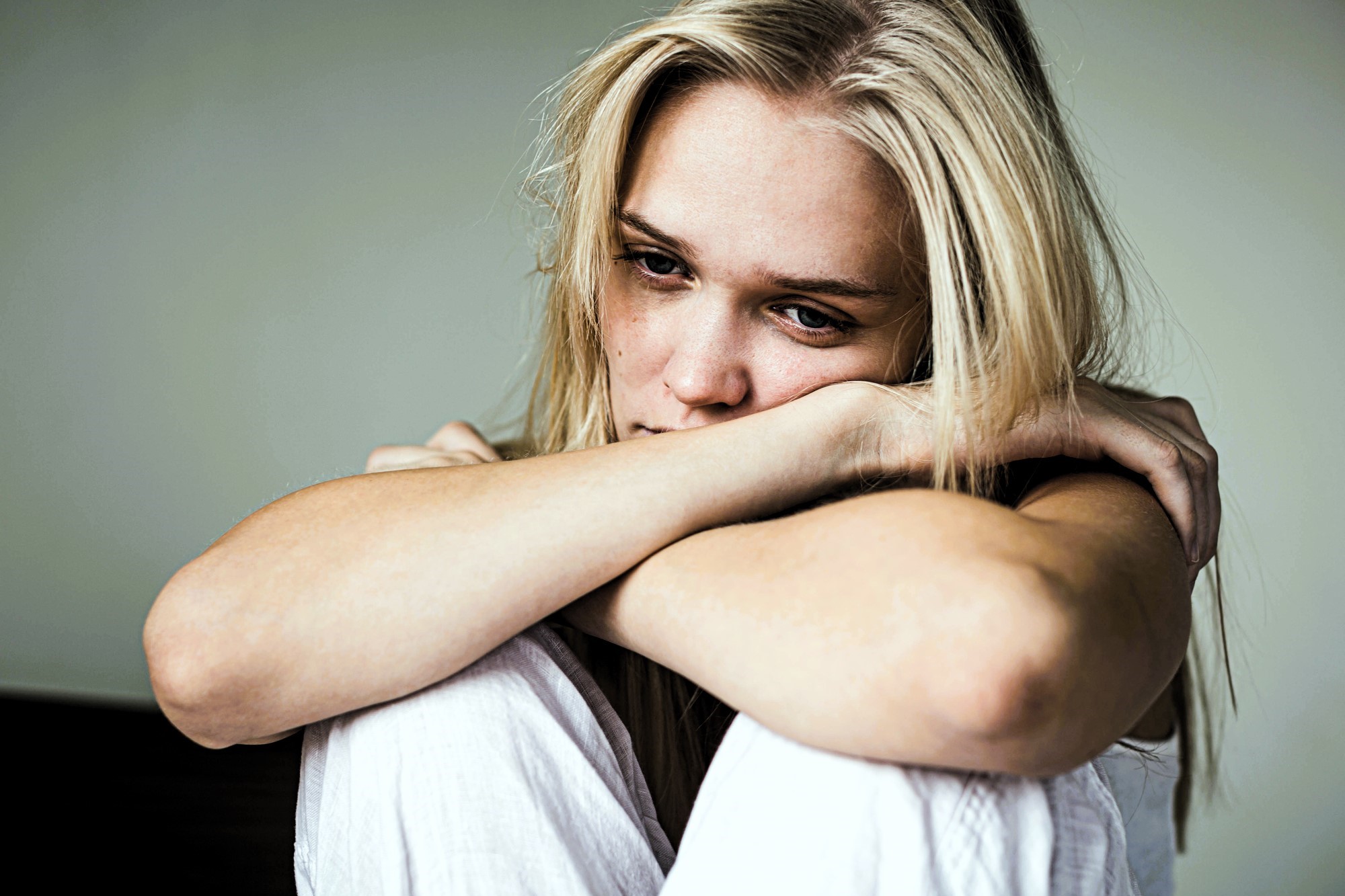 A woman with long blonde hair sits with her arms resting on her knees, looking contemplatively into the distance. She appears to be in a relaxed indoor setting, wearing a white outfit. The background is blurred.