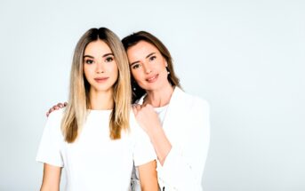 Two women with long hair stand closely together against a light background. One woman rests her hand on the other's shoulder. Both are wearing white tops and smiling softly at the camera.