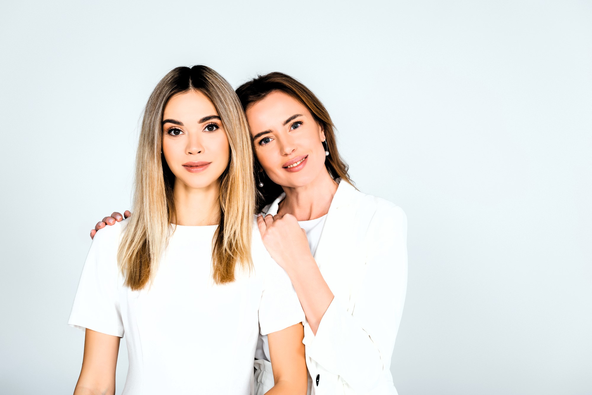 Two women with long hair stand closely together against a light background. One woman rests her hand on the other's shoulder. Both are wearing white tops and smiling softly at the camera.