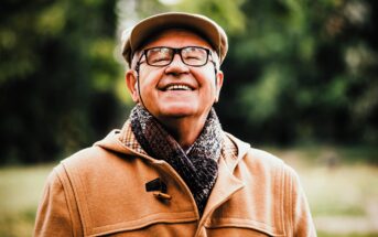 An elderly man smiles broadly while standing outdoors. He wears glasses, a beige flat cap, a brown coat, and a dark patterned scarf. The background is blurred greenery, suggesting a park or garden setting.