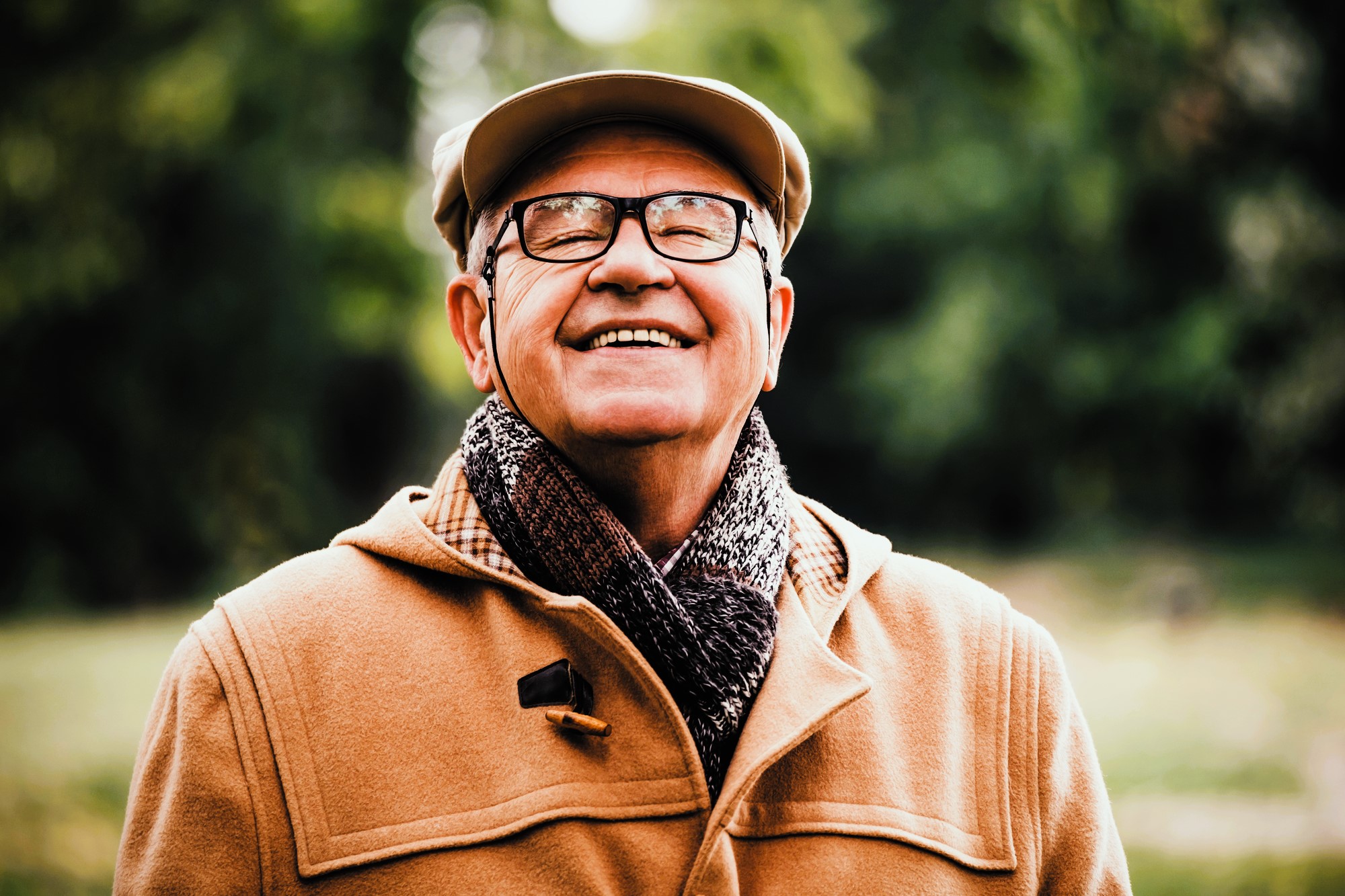 An elderly man smiles broadly while standing outdoors. He wears glasses, a beige flat cap, a brown coat, and a dark patterned scarf. The background is blurred greenery, suggesting a park or garden setting.