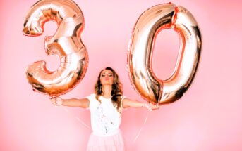 Woman in a white shirt and pink skirt holds large rose gold balloons shaped as the number "30" against a pink background. She is puckering her lips and standing with arms outstretched.