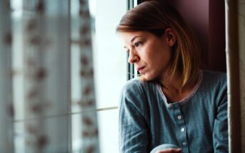 A woman with shoulder-length hair wearing a blue long-sleeve shirt looks pensively out a window. She sits near sheer curtains, with soft light illuminating her thoughtful expression.