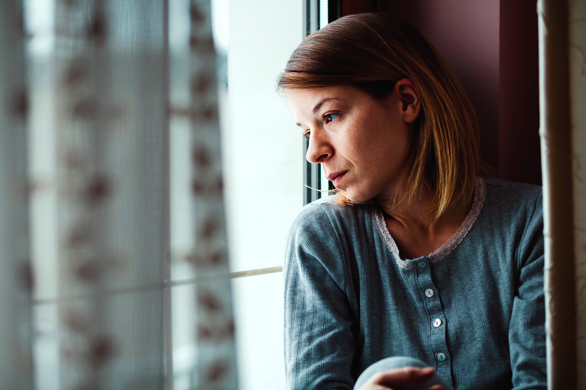 A woman with shoulder-length hair wearing a blue long-sleeve shirt looks pensively out a window. She sits near sheer curtains, with soft light illuminating her thoughtful expression.