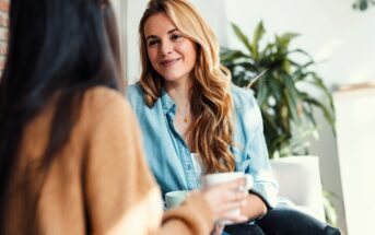 Two people sitting indoors, engaged in conversation. One person with long blonde hair in a denim shirt smiles while holding a mug. The other is seen from behind. A plant and natural light are in the background.