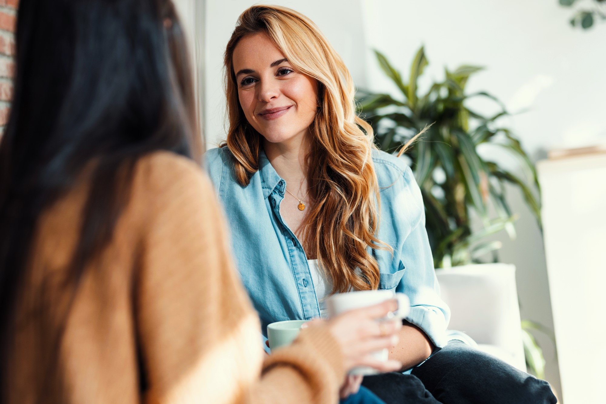 Two people sitting indoors, engaged in conversation. One person with long blonde hair in a denim shirt smiles while holding a mug. The other is seen from behind. A plant and natural light are in the background.