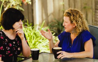 Two women sitting at a patio table engaged in a conversation. One has dark, shoulder-length hair and wears a floral dress, while the other has curly blonde hair and wears a blue blouse. They each have a black mug in front of them.