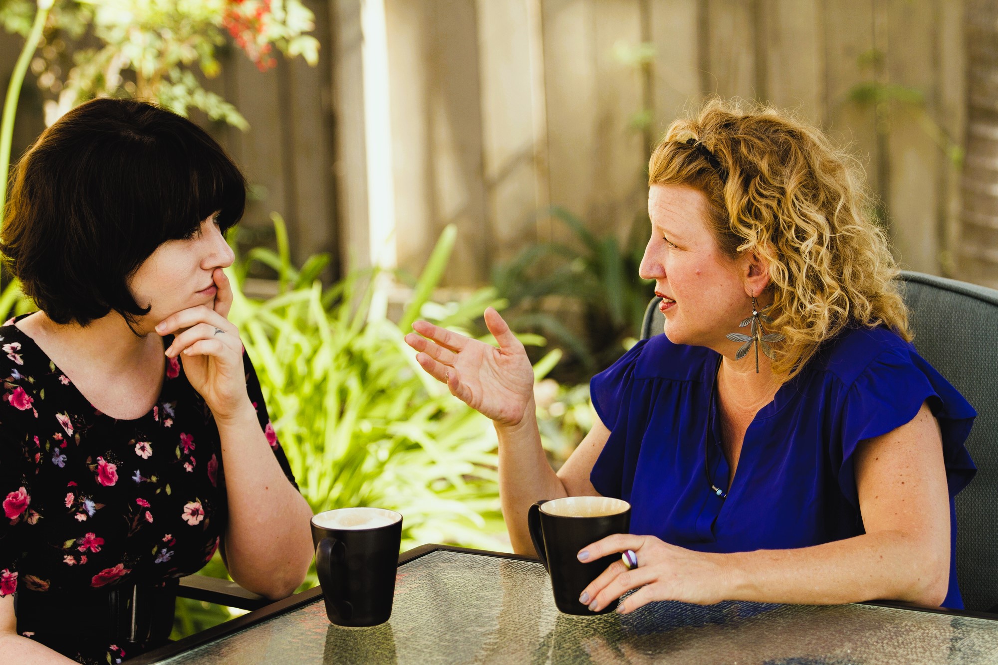 Two women sitting at a patio table engaged in a conversation. One has dark, shoulder-length hair and wears a floral dress, while the other has curly blonde hair and wears a blue blouse. They each have a black mug in front of them.