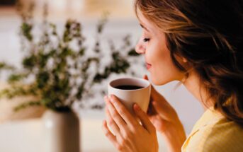 A woman in a yellow top is sitting with her eyes closed, holding a cup of coffee near her face. She appears to be inhaling its aroma. In the background, there is a blurred plant in a vase.