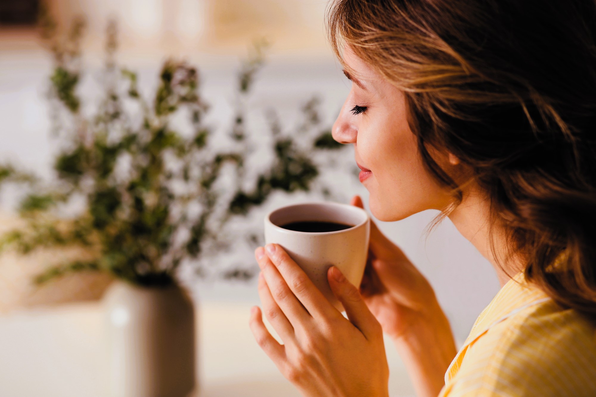 A woman in a yellow top is sitting with her eyes closed, holding a cup of coffee near her face. She appears to be inhaling its aroma. In the background, there is a blurred plant in a vase.