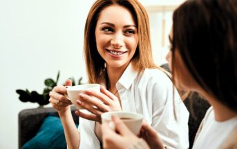 Two women sitting on a couch are smiling and holding coffee mugs. One woman has red hair and is looking at the other, who has brown hair. There are green plants and a blue pillow in the background.