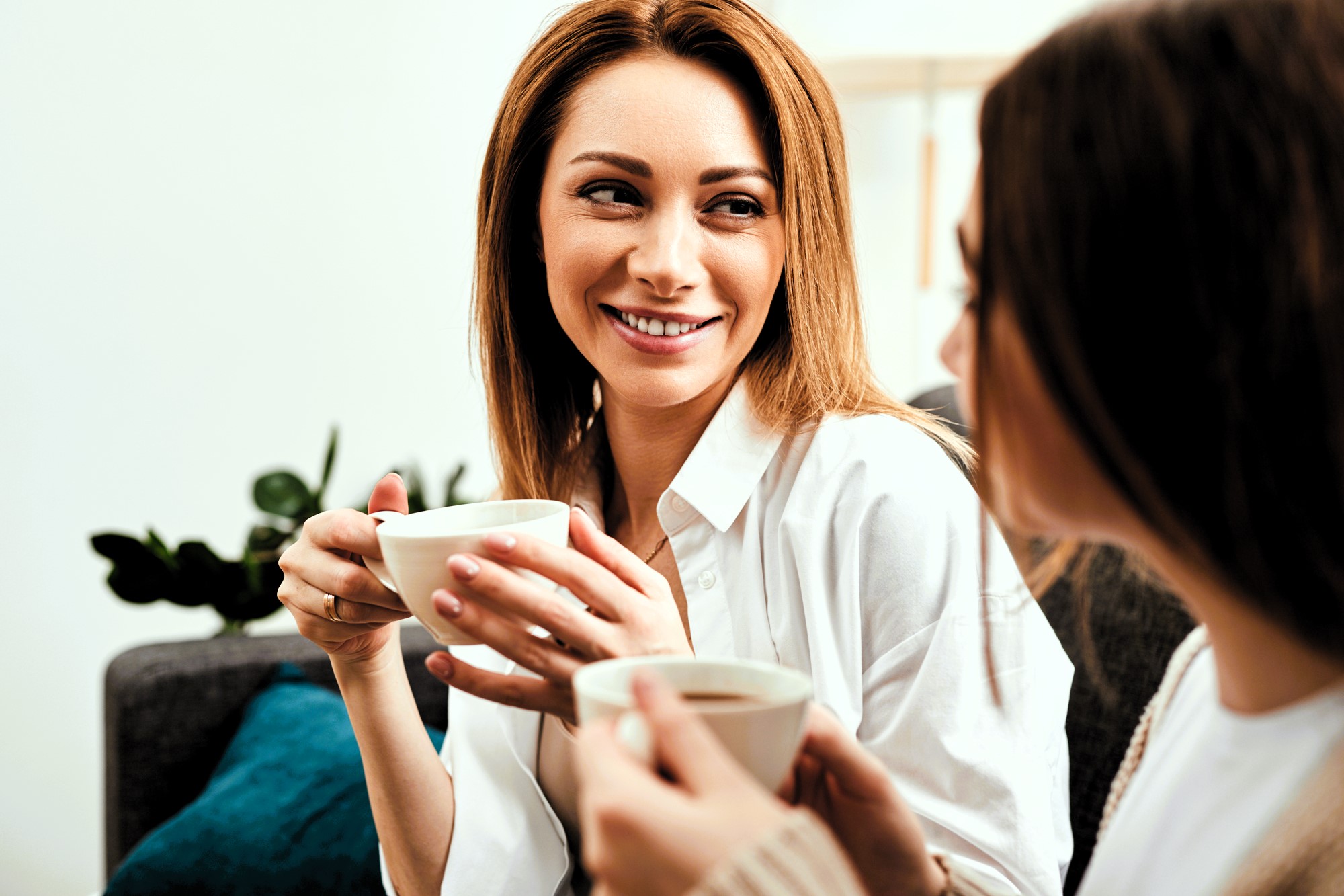 Two women sitting on a couch are smiling and holding coffee mugs. One woman has red hair and is looking at the other, who has brown hair. There are green plants and a blue pillow in the background.