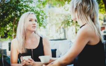 Two people with light hair sit outdoors on a patio, holding white mugs. One is facing forward, listening attentively, while the other is turned sideways, speaking. They are surrounded by greenery and a soft, natural light.