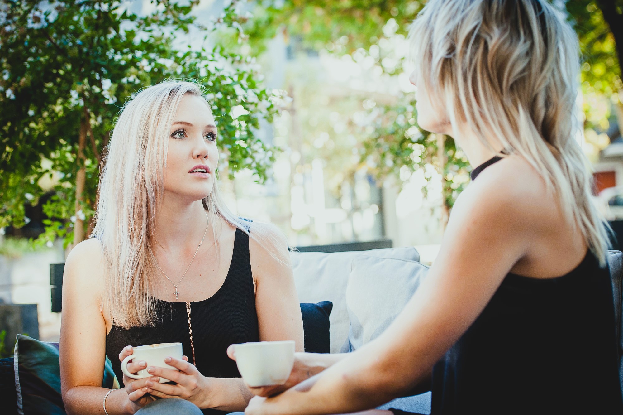 Two people with light hair sit outdoors on a patio, holding white mugs. One is facing forward, listening attentively, while the other is turned sideways, speaking. They are surrounded by greenery and a soft, natural light.