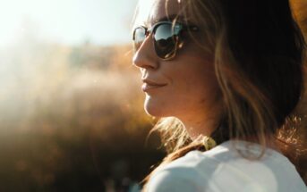 A woman with long hair is wearing sunglasses, looking towards the sunlight. She is outdoors, with a blurred background of trees and a bright sky. The light creates a soft, warm glow on her face.