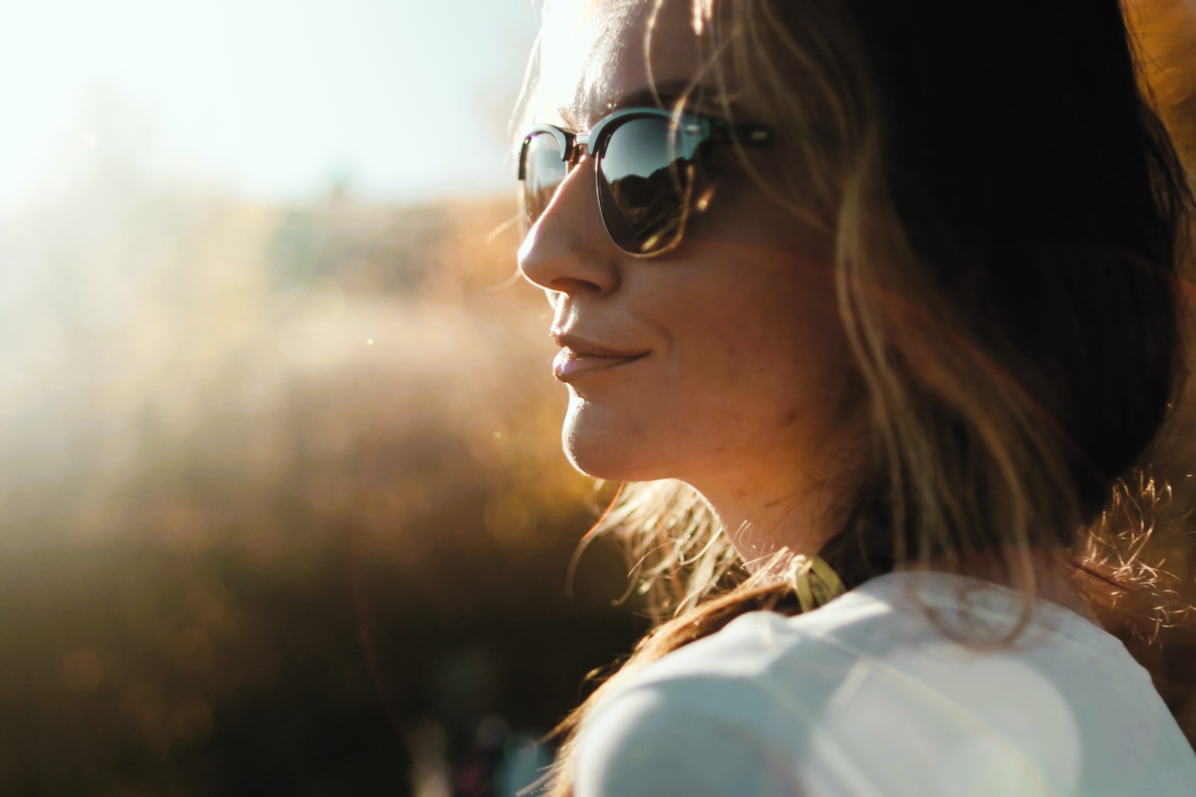 A woman with long hair is wearing sunglasses, looking towards the sunlight. She is outdoors, with a blurred background of trees and a bright sky. The light creates a soft, warm glow on her face.