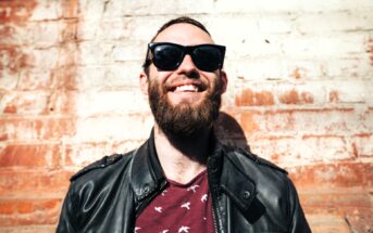 A bearded man wearing sunglasses and a leather jacket smiles broadly while standing against a textured brick wall. The sunlight casts a warm glow on the scene.