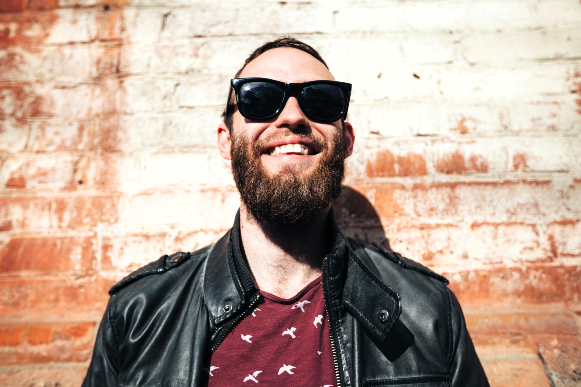 A bearded man wearing sunglasses and a leather jacket smiles broadly while standing against a textured brick wall. The sunlight casts a warm glow on the scene.