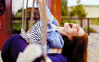 A woman with long brown hair laughs joyfully while sitting on a swing. She wears a striped shirt and blue jeans. Her eyes are closed, and she is leaning back, grasping the swing chains, with a park setting in the background.