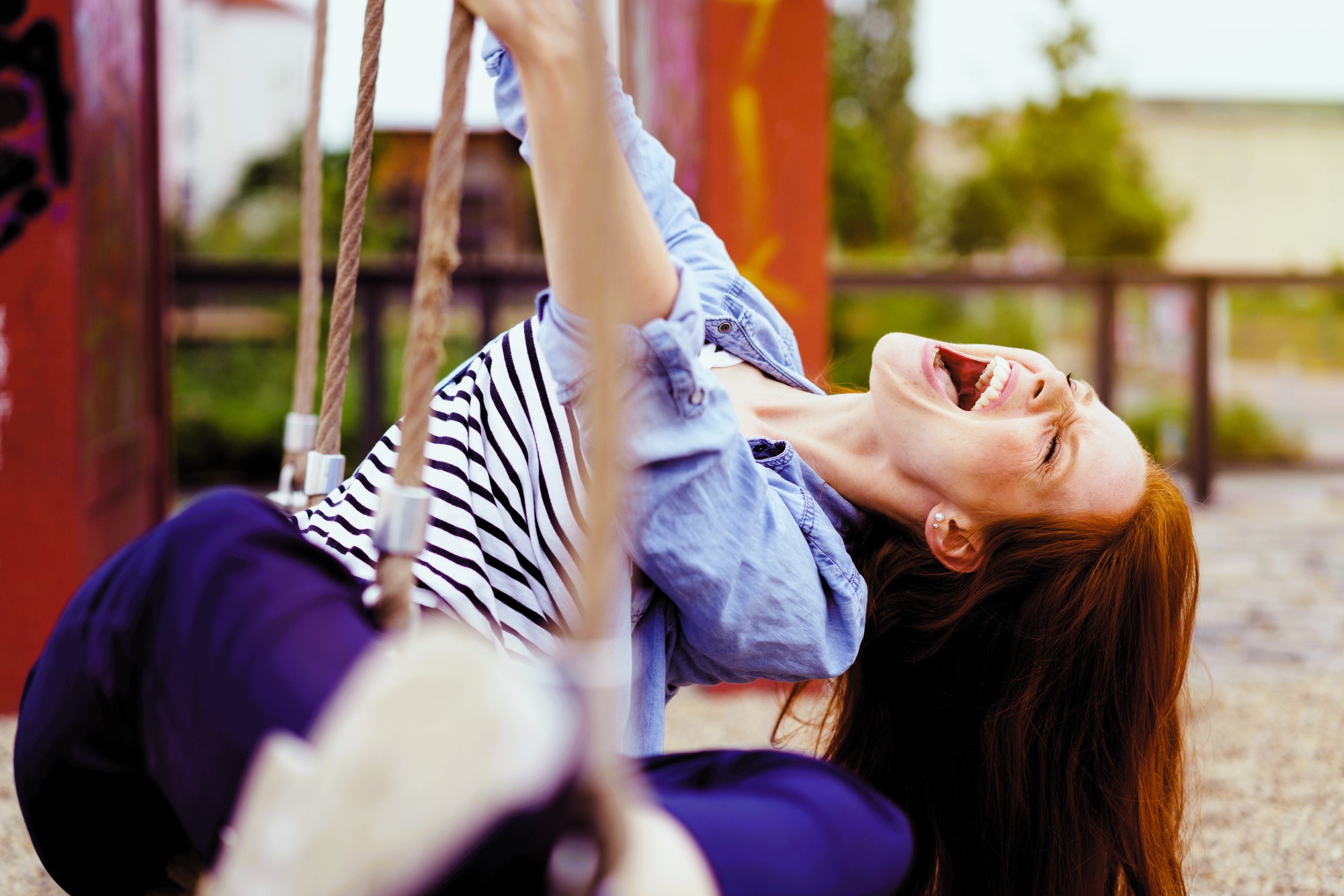 A woman with long brown hair laughs joyfully while sitting on a swing. She wears a striped shirt and blue jeans. Her eyes are closed, and she is leaning back, grasping the swing chains, with a park setting in the background.