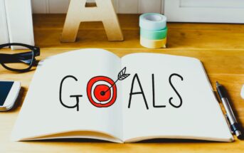 An open notebook on a wooden table with the word "Goals" written on it. The "O" is stylized as a red target with an arrow. Nearby are glasses, a smartphone, tape rolls, and a pen.