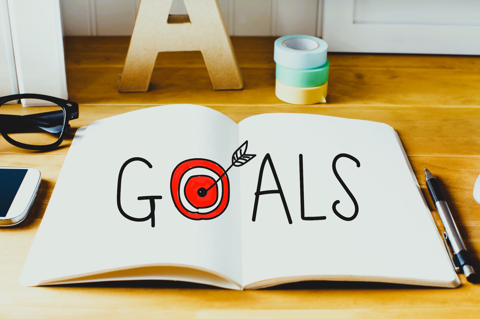 An open notebook on a wooden table with the word "Goals" written on it. The "O" is stylized as a red target with an arrow. Nearby are glasses, a smartphone, tape rolls, and a pen.