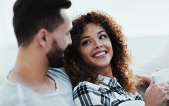 A man and woman sit together on a couch, drinking from white mugs. The woman, with curly hair in a plaid shirt, looks at the man and smiles. The man, with short hair and a beard, wears a gray shirt and looks at the woman. They appear relaxed and happy.