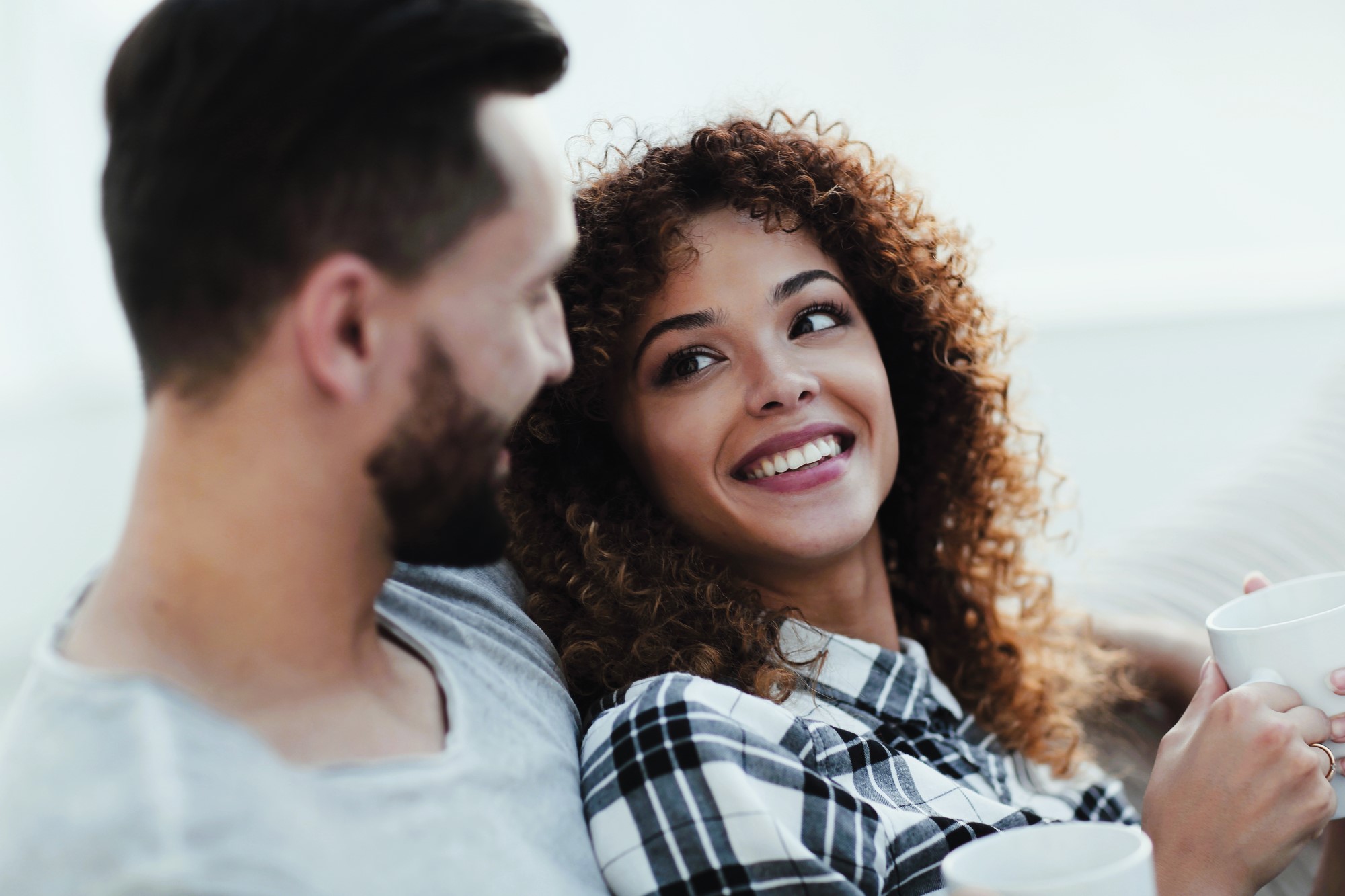 A man and woman sit together on a couch, drinking from white mugs. The woman, with curly hair in a plaid shirt, looks at the man and smiles. The man, with short hair and a beard, wears a gray shirt and looks at the woman. They appear relaxed and happy.