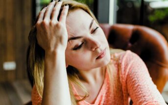 A woman with long blonde hair, wearing a peach-colored sweater, rests her head on her hand while sitting on a brown leather chair. She appears thoughtful and looks off to the side, with a blurred background of a room.