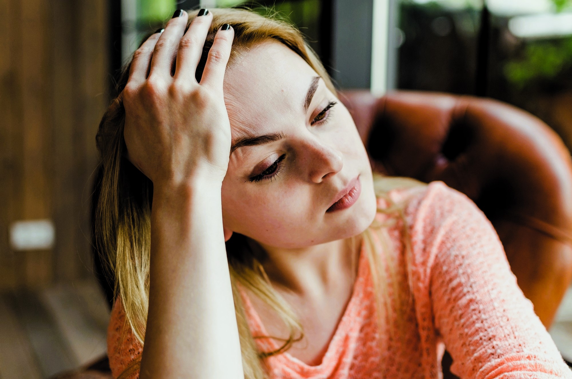 A woman with long blonde hair, wearing a peach-colored sweater, rests her head on her hand while sitting on a brown leather chair. She appears thoughtful and looks off to the side, with a blurred background of a room.