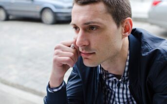 A man with short brown hair is deep in thought, resting his chin on his hand. He is wearing a navy jacket over a checkered shirt. Parked cars can be seen in the blurred background on the street.