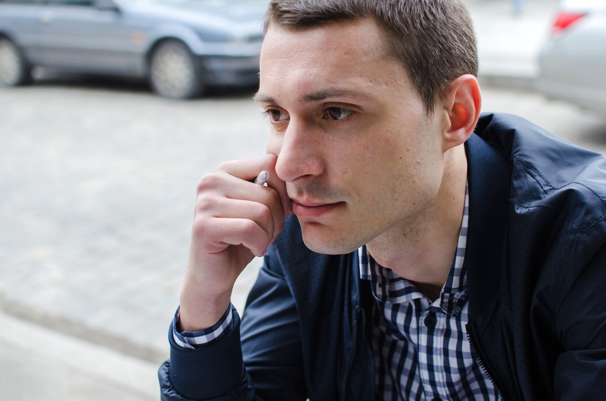 A man with short brown hair is deep in thought, resting his chin on his hand. He is wearing a navy jacket over a checkered shirt. Parked cars can be seen in the blurred background on the street.