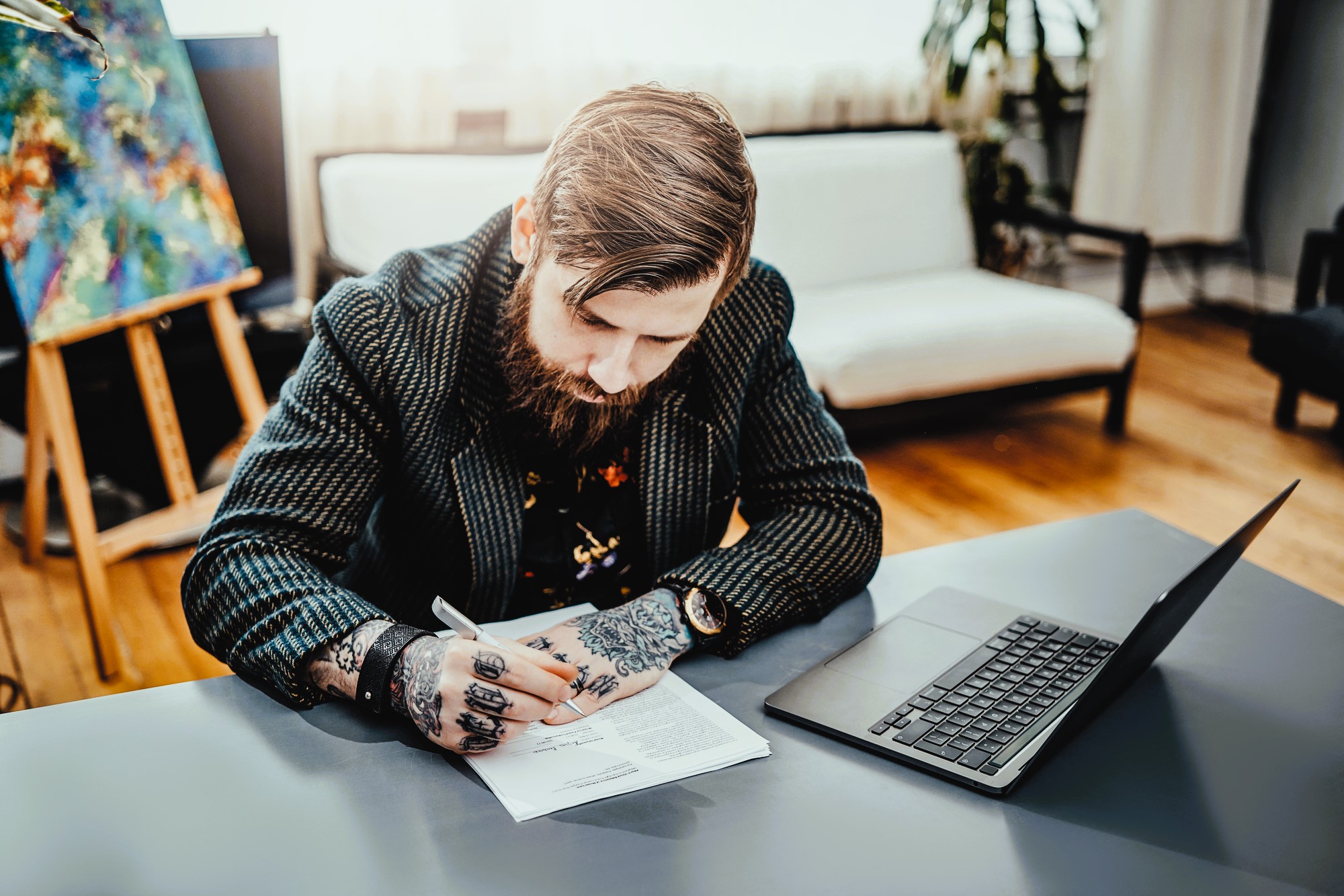 A bearded man with tattoos on his hands is seated at a desk, writing on a document. A laptop is open beside him. The background features a couch, a large window, and a colorful painting on an easel.