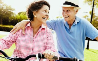 An older couple happily riding bicycles in a sunny park. The woman wears a pink checkered shirt, and the man wears a blue polo and a fedora. They're smiling and looking at each other, with trees and grass in the background.