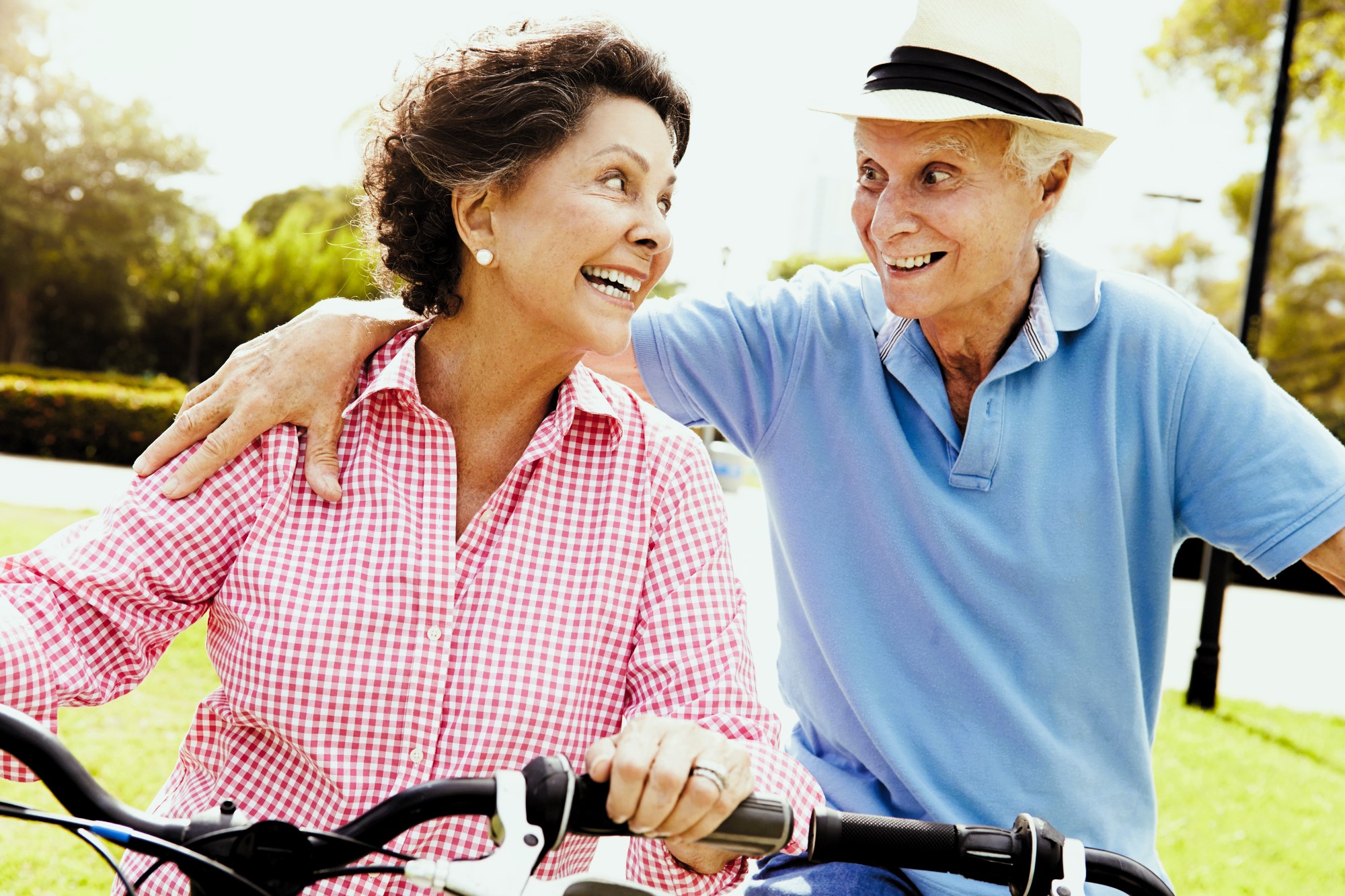 An older couple happily riding bicycles in a sunny park. The woman wears a pink checkered shirt, and the man wears a blue polo and a fedora. They're smiling and looking at each other, with trees and grass in the background.