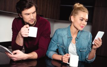 A man with a maroon shirt holds a coffee mug and newspaper, glancing sideways. A woman in a denim jacket sits beside him, holding a mug and looking at her phone. They are in a modern kitchen with dark cabinets.