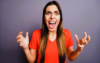 A woman in a red shirt stands against a gray background, expressing frustration or anger. Her mouth is open wide, eyes are wide, and hands are raised with fingers spread apart, as if yelling or exclaiming.