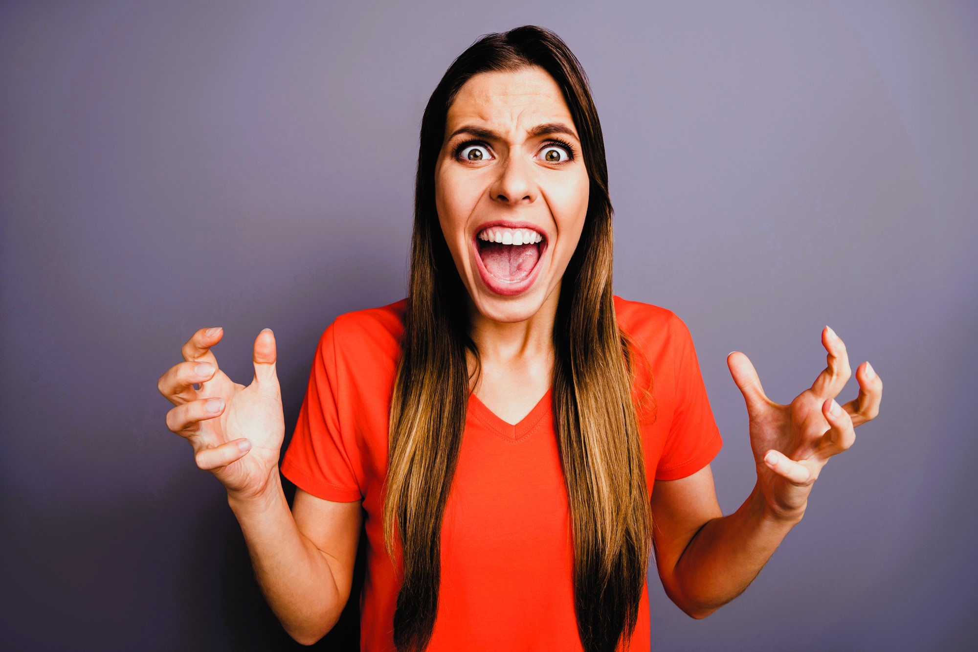 A woman in a red shirt stands against a gray background, expressing frustration or anger. Her mouth is open wide, eyes are wide, and hands are raised with fingers spread apart, as if yelling or exclaiming.