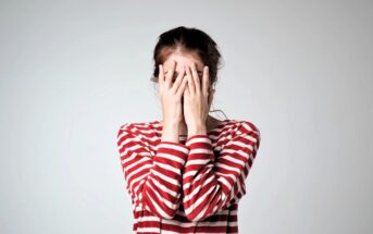 A person wearing a red and white striped shirt covers their face with both hands. The background is light gray, creating a simple and neutral setting.