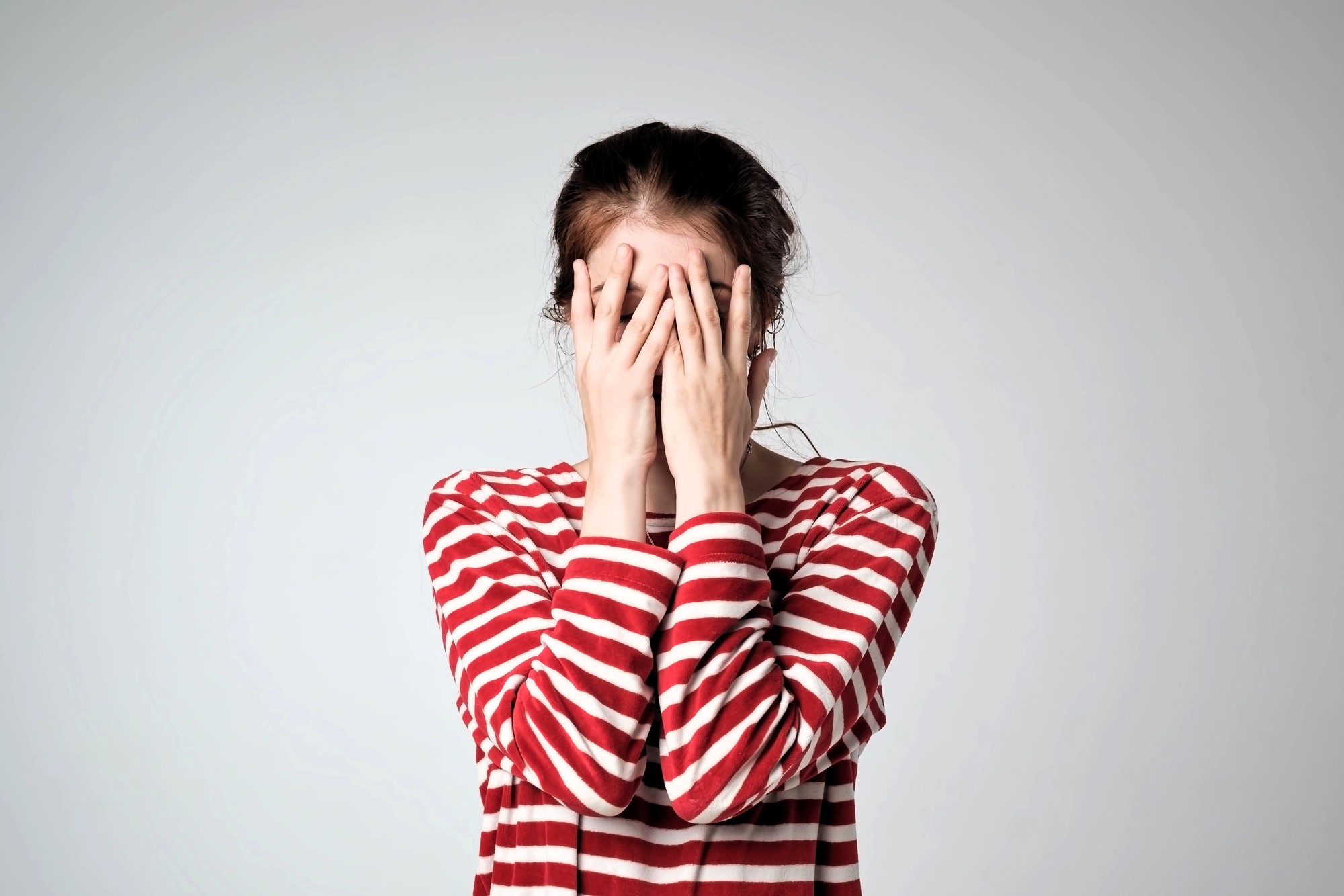 A person wearing a red and white striped shirt covers their face with both hands. The background is light gray, creating a simple and neutral setting.