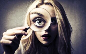 A woman with long blonde hair looks closely through a magnifying glass, which enlarges one of her eyes. The background is a plain, textured wall. Her expression is curious and slightly dramatic.